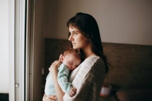 Mother holding her sleeping baby against her chest while looking out the window with a tired, reflective expression. Soft natural light falls on them, while the background remains dim, capturing a quiet moment of maternal exhaustion, love, and emotional overwhelm.
